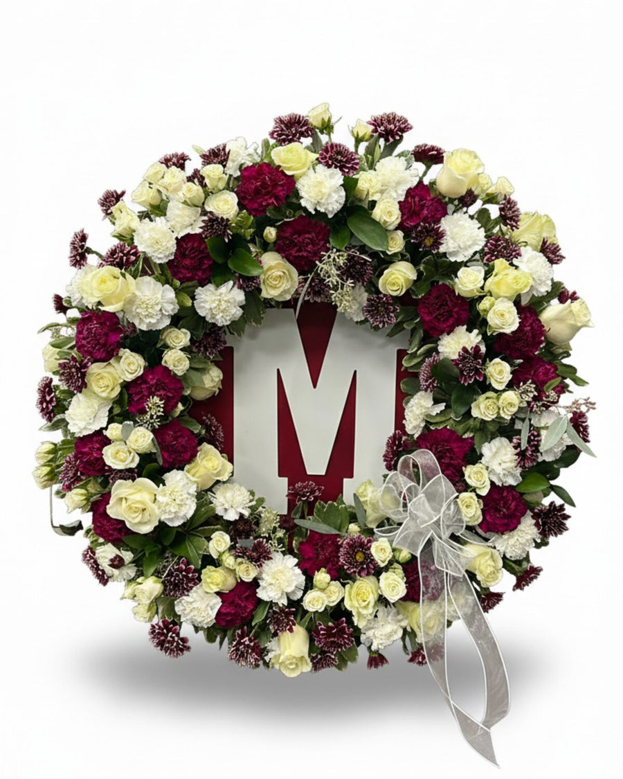 Monogram Wreath — a round standing wreath of burgundy and cream carnations with cream roses, finished with a silver ribbon and a single letter in the center.