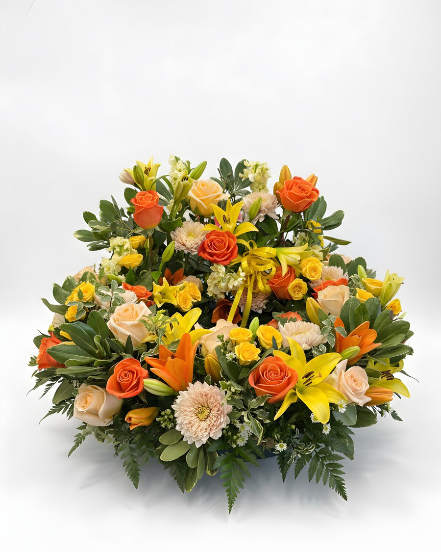 Golden Tribute — a full, rounded arrangement of orange and peach roses with yellow and orange lilies, peach mums, fern and fresh greenery.