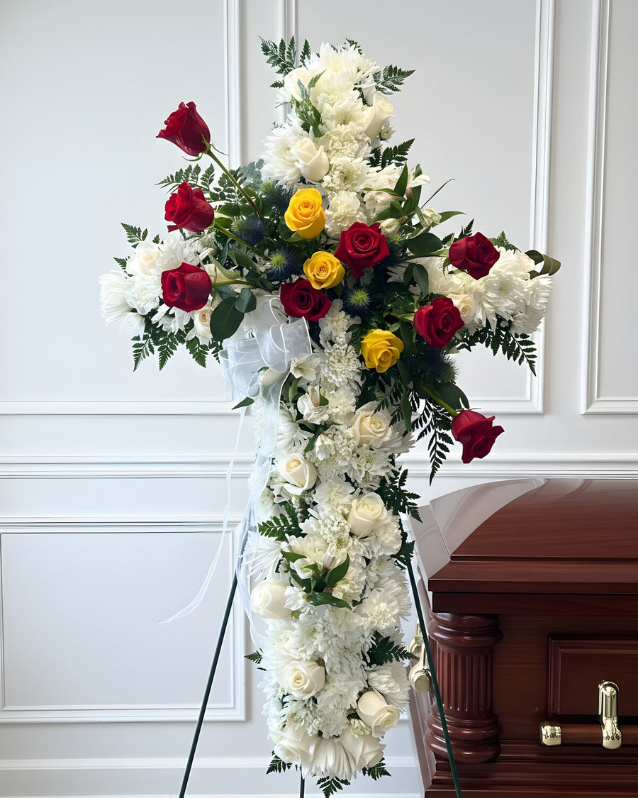 Standing Cross — a standing cross of white carnations accented with red and yellow roses and fresh greenery, finished with a white ribbon and displayed on an easel.