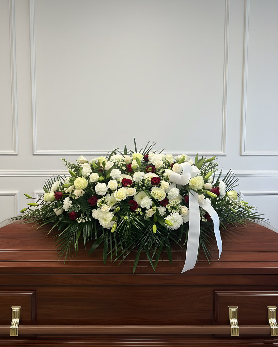 Peaceful White Casket Spray — a casket spray of white and cream roses and white carnations, with a few deep red roses and fresh palm and fern, finished with a white ribbon.