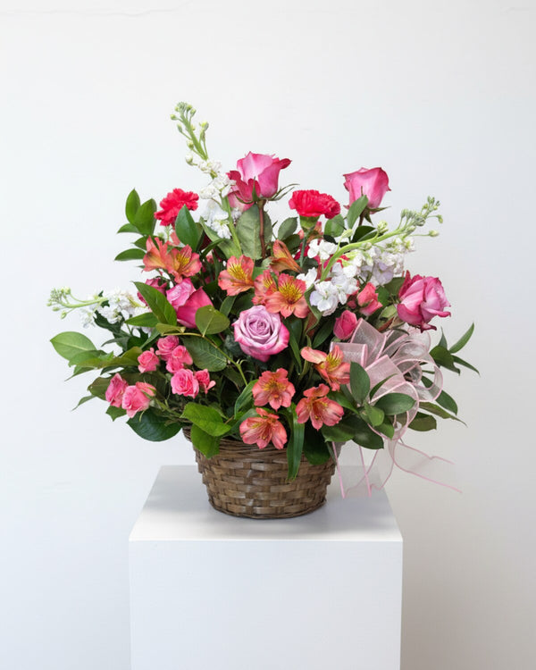 Coral — a woven basket of pink and lavender roses with coral alstroemeria, white stock and deep pink spray roses, finished with a pink ribbon.