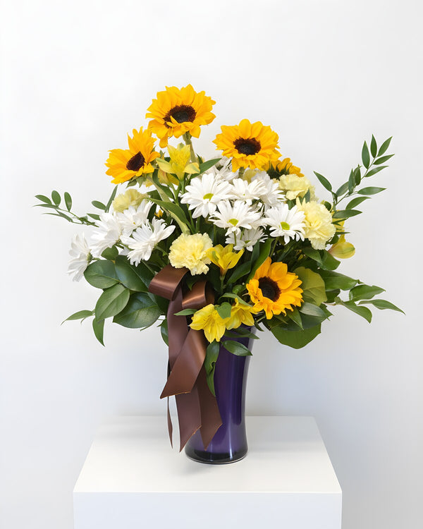 Sunny Side — sunflowers, white daisies and yellow carnations in a purple vase with a bronze ribbon