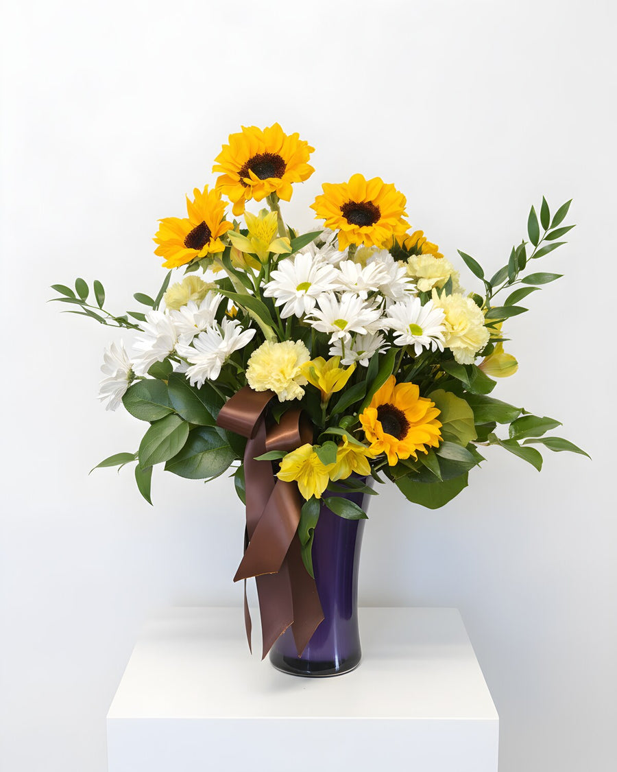 Sunny Side — sunflowers, white daisies and yellow carnations in a purple vase with a bronze ribbon