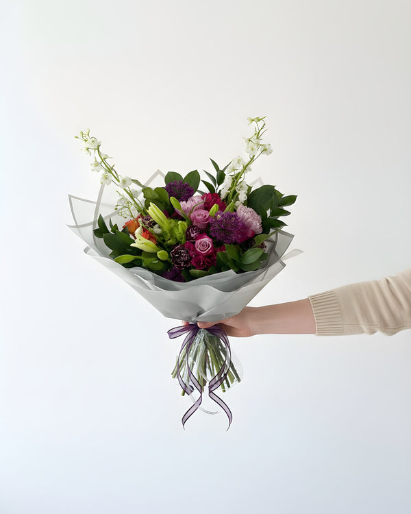Lavender Field — a hand-tied bouquet of purple and lavender blooms with white stock, lily buds and fresh greens, wrapped in white paper and tied with a purple ribbon.