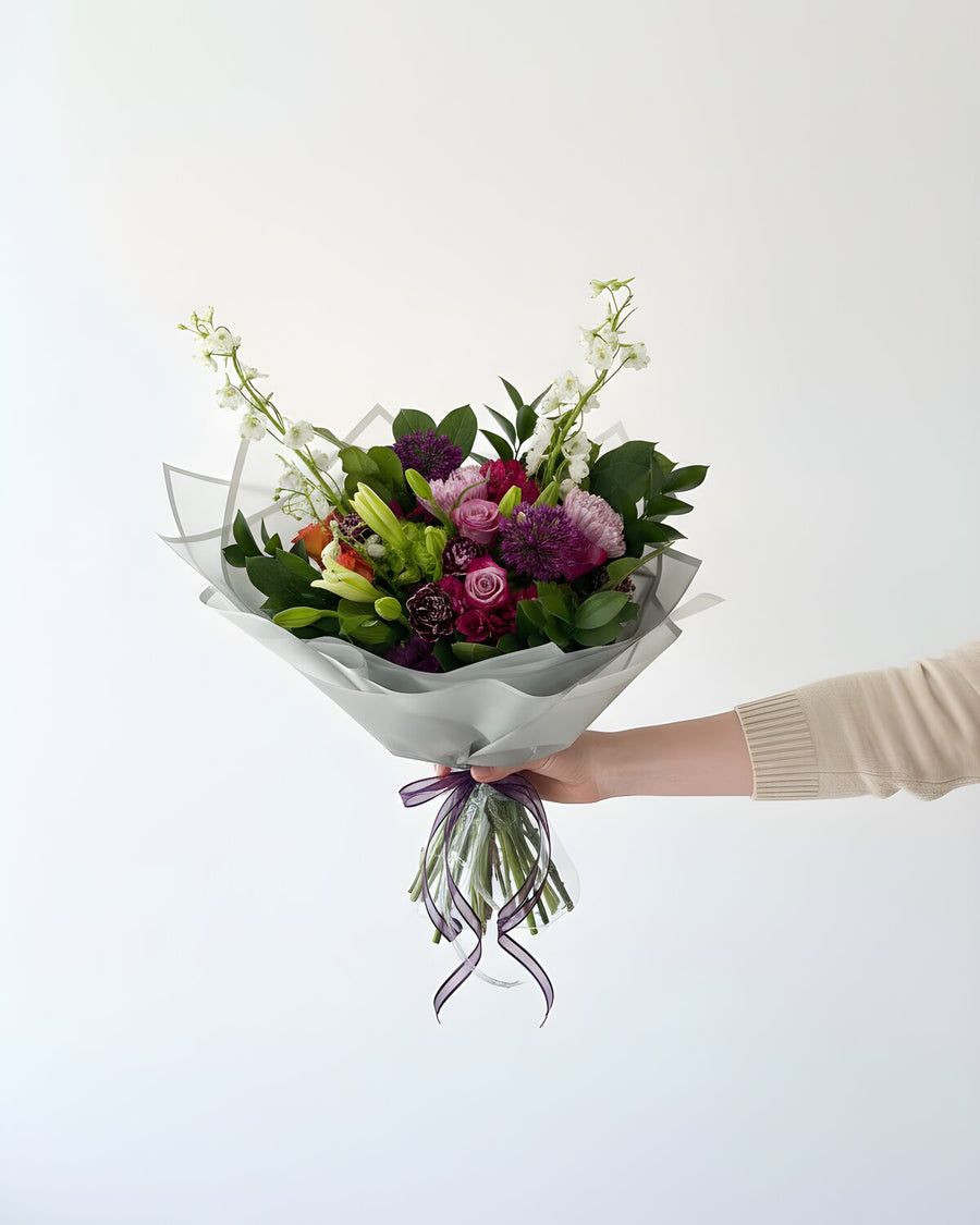Lavender Field — a hand-tied bouquet of purple and lavender blooms with white stock, lily buds and fresh greens, wrapped in white paper and tied with a purple ribbon.
