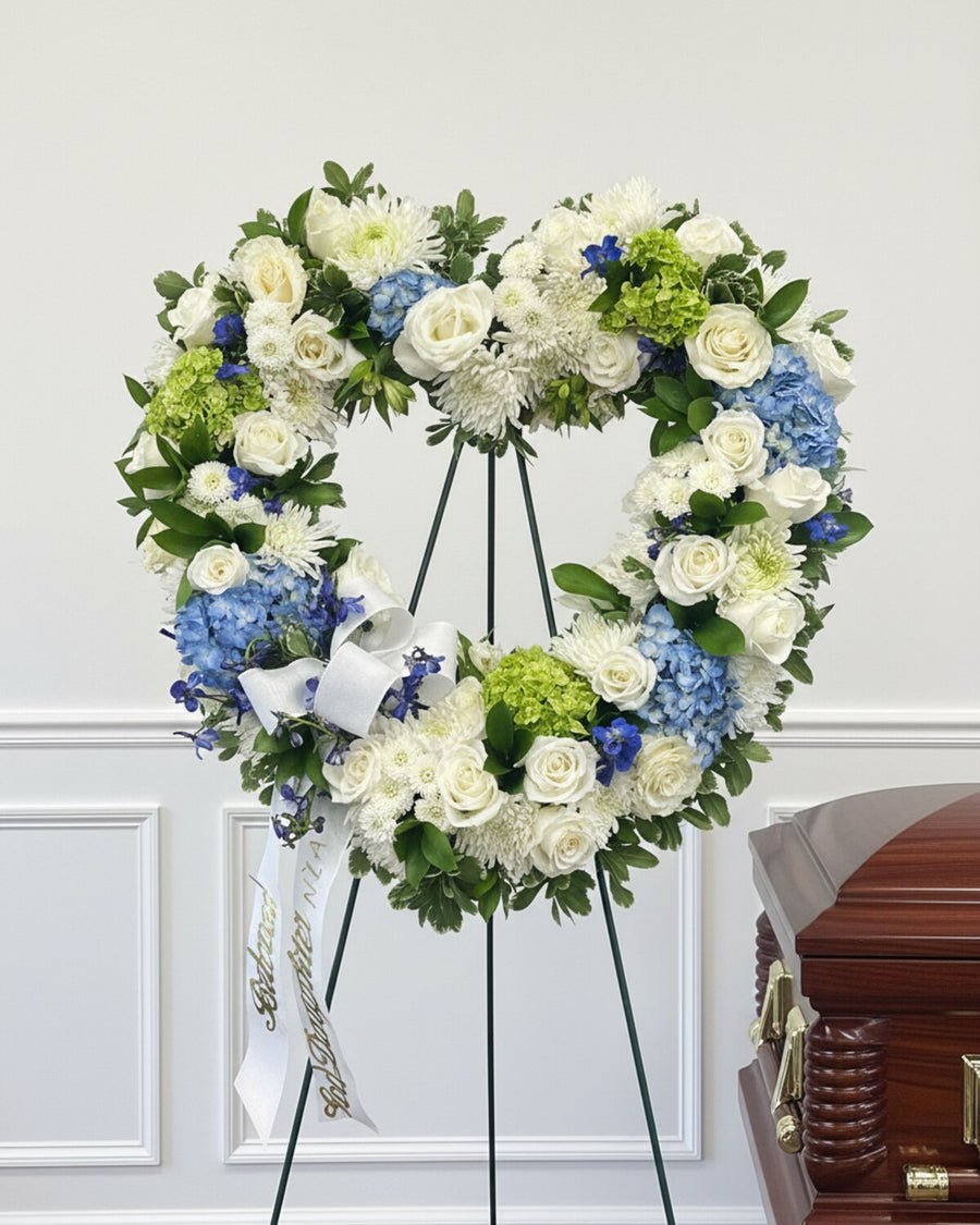 Serenity Wreath — a heart-shaped standing wreath of white roses with blue and green hydrangea, finished with a white ribbon and displayed on an easel.