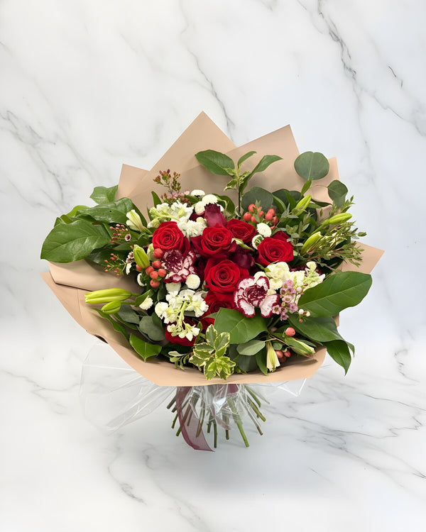Winter Wrap — a wrapped bouquet of red roses with pink and white carnations, white lilies, red hypericum berries and fresh greens, finished in soft blush paper.