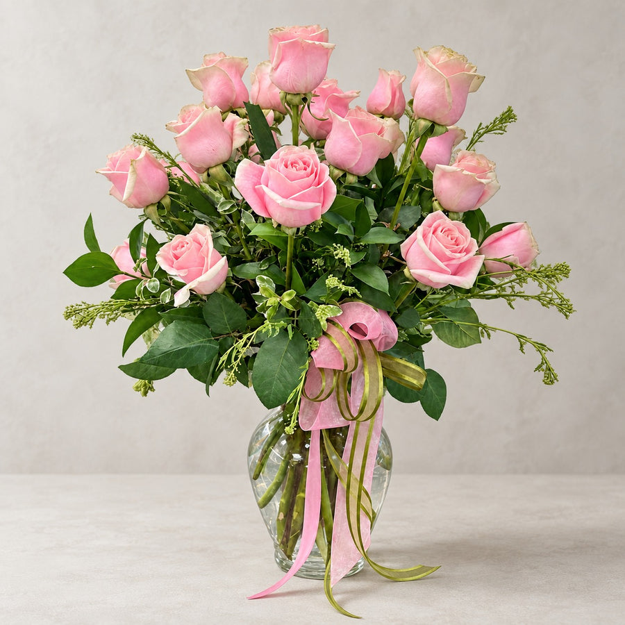 Blush Dozen — soft pink roses with fresh greenery in a clear glass vase, tied with a pink ribbon