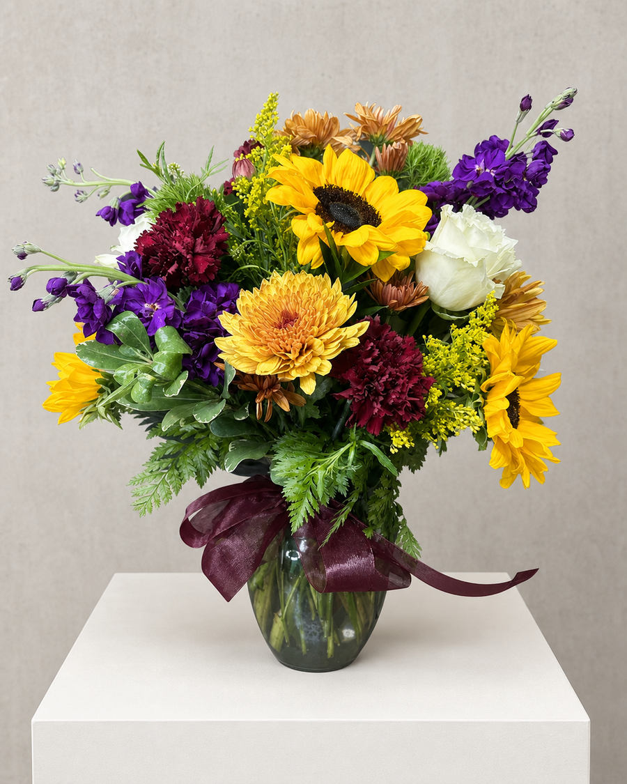 Late Light — a bold, sunny arrangement in a glass vase: sunflowers and golden mums with deep burgundy carnations, purple stock, a white rose and bright solidago, finished with a burgundy ribbon.
