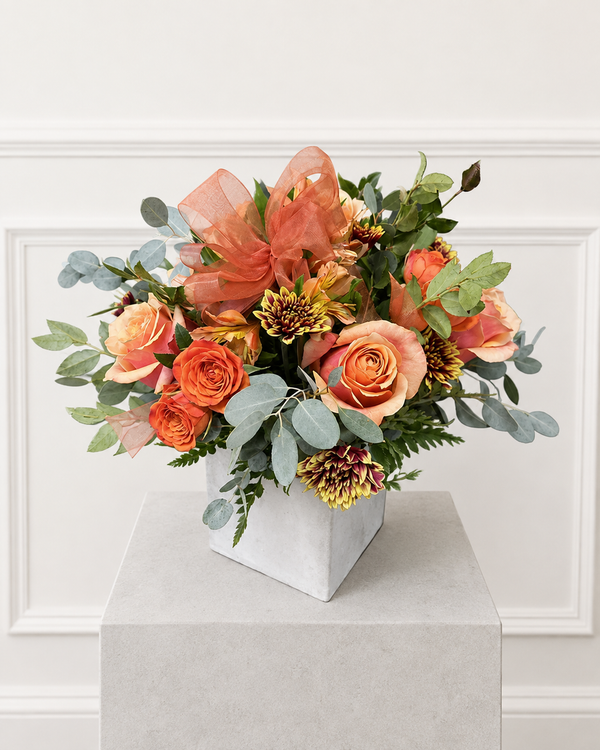 October Blush — a fall arrangement of orange and peach roses with bronze mums and silver-green eucalyptus in a white ceramic cube, finished with a peach bow.