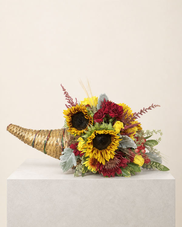 The Long Table — a woven cornucopia overflowing with sunflowers, deep red blooms, dusty miller, wheat and fall accents.